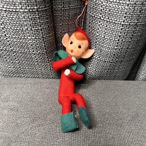 Può includere: Un ornamento natalizio vintage a forma di elfo. L'elfo ha un corpo in feltro rosso, un cappello rosso e colletto e stivali verdi. Il viso dell'elfo è dipinto con guance rosee e un'espressione dolce. Un piccolo anello di spago è attaccato per appenderlo.