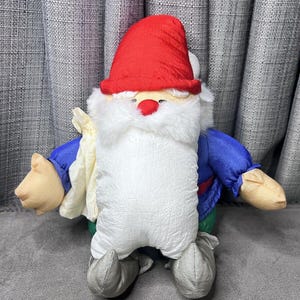 May include: A plush gnome toy with a red hat, white beard, blue shirt, and green trousers. The gnome is holding a white bag in its left hand.