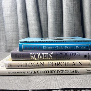 May include: A stack of five books about porcelain and pottery. The top book is blue and says "1100 Marks on Foreign Pottery & Porcelain". The next book is blue and says "Kovels' Dictionary of Marks-Pottery & Porcelain". The third book is purple and says "Kovels' New Dictionary of Marks". The fourth book is cream and says "German Porcelain". The bottom book is white and says "The Color Treasury of 18th Century Porcelain".