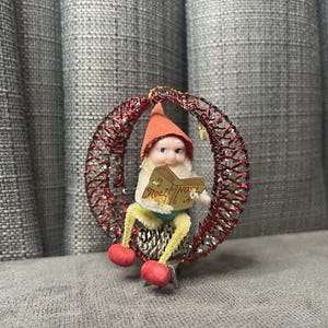 May include: A vintage Christmas ornament featuring a small elf figure with a red hat and holding a book that says "Noel". The elf is seated on a pinecone base within a red wire wreath. The ornament has red and gold accents.
