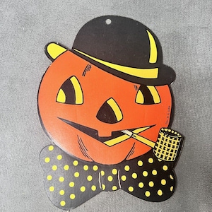 Könnte beinhalten: Vintage Halloween-Dekoration mit einem orangefarbenen Kürbis mit schwarzem Hut und Fliege. Der Kürbis hat gelbe Augen und einen lächelnden Mund, der eine Pfeife hält. Die Fliege ist schwarz mit gelben Punkten. Ein klassisches Halloween-Design.