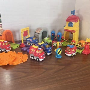 May include: A colorful toy train set with various components, including a red fire truck, blue and green trains, a tunnel, a station, and track pieces. The set features orange, red, yellow, and blue elements, designed for imaginative play.