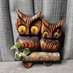 May include: Two wooden owl figurines with detailed carved features and yellow eyes. The owls are perched on a wooden log with small green leaves. The wood has a rich brown color with visible grain.