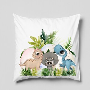 May include: A white pillow with a colorful illustration of three dinosaurs in a jungle setting. The dinosaurs are a blue dinosaur, a brown dinosaur, and a gray dinosaur. The dinosaurs are surrounded by green leaves and pink, blue, and white eggs.