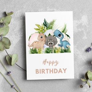 May include: A white card with a watercolor illustration of three dinosaurs in a nest of green leaves. The dinosaurs are a blue dinosaur, a brown dinosaur, and a gray dinosaur. The text "Happy Birthday" is printed in brown on the card.