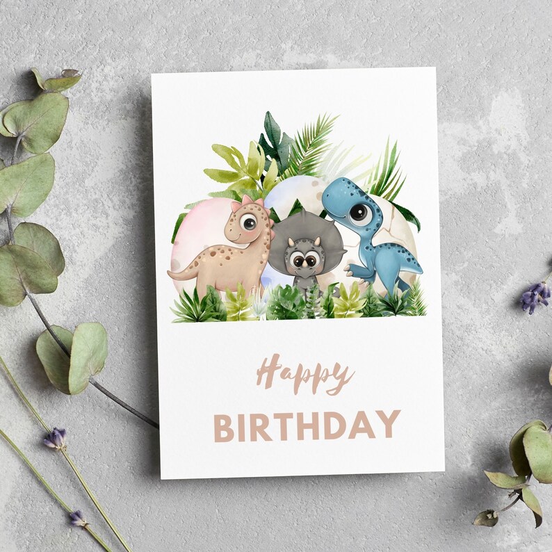May include: A white card with a watercolor illustration of three dinosaurs in a nest of green leaves. The dinosaurs are a blue dinosaur, a brown dinosaur, and a gray dinosaur. The text "Happy Birthday" is printed in brown on the card.