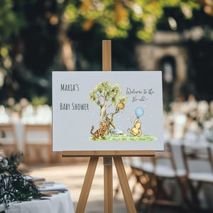 May include: A light gray baby shower sign on a wooden easel. The sign has the text "Maria's Baby Shower" and "Welcome to the World". It features illustrations of a tree, a tiger, and a bear holding a blue balloon.