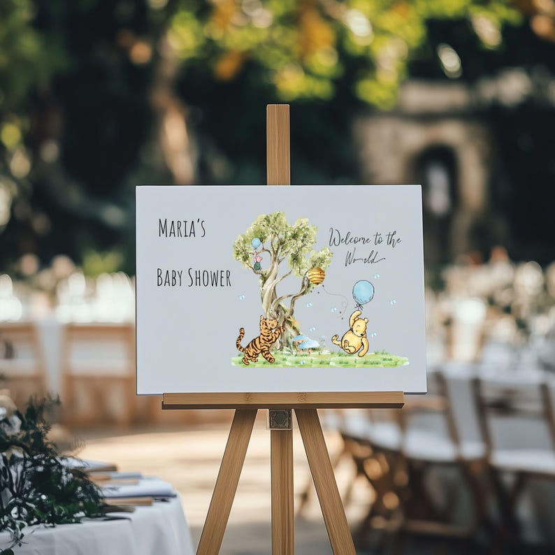May include: A light gray baby shower sign on a wooden easel. The sign has the text "Maria's Baby Shower" and "Welcome to the World". It features illustrations of a tree, a tiger, and a bear holding a blue balloon.