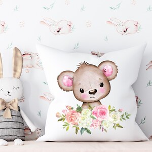 May include: White pillow with a brown bear face surrounded by pink and white flowers. The bear has pink ears and a brown nose. The background is white with a repeating pattern of pink bunnies.