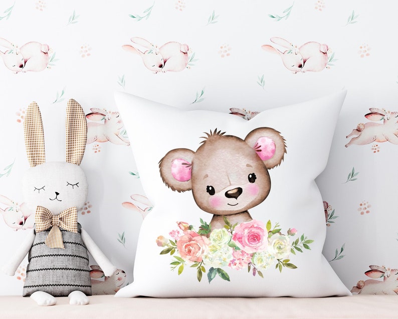 May include: White pillow with a brown bear face surrounded by pink and white flowers. The bear has pink ears and a brown nose. The background is white with a repeating pattern of pink bunnies.