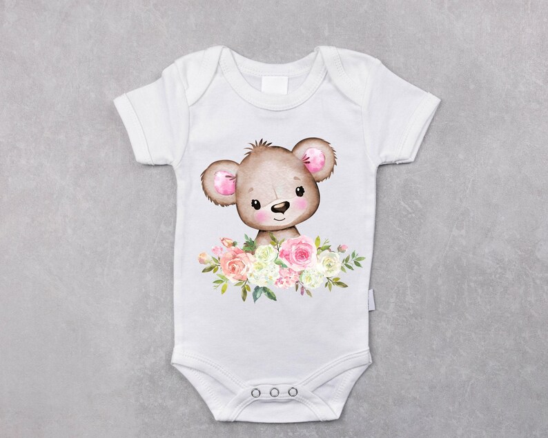 May include: White baby onesie with a brown bear with pink ears and a floral design in shades of pink, white, and green.
