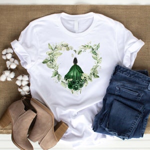 K&ouml;nnte beinhalten: Wei&szlig;es T-Shirt mit einem gr&uuml;nen floralen Herzdesign, das eine Frau in einem gr&uuml;nen Kleid zeigt. Das Design ist mittig auf dem Shirt platziert.