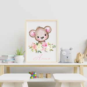May include: A framed print featuring a watercolor illustration of a brown bear with pink ears and a pink nose. The bear is surrounded by pink and white flowers. The name "Anna" is written in pink cursive below the bear.
