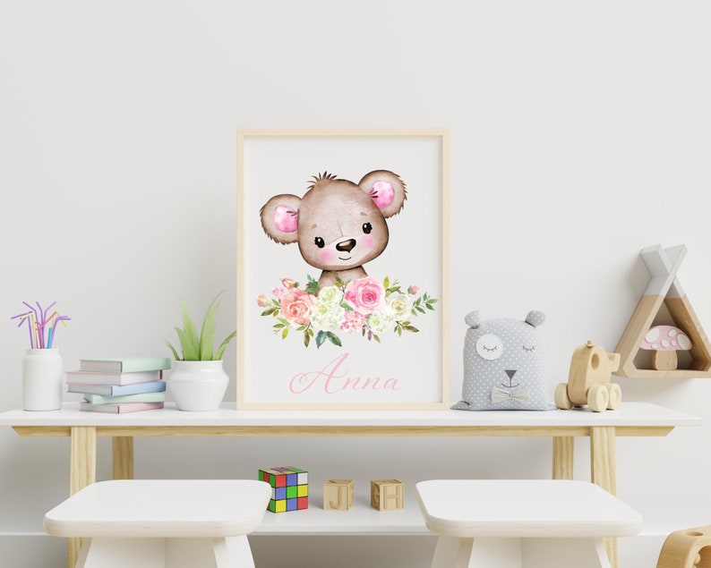 May include: A framed print featuring a watercolor illustration of a brown bear with pink ears and a pink nose. The bear is surrounded by pink and white flowers. The name "Anna" is written in pink cursive below the bear.