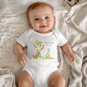 May include: A white baby onesie featuring a colorful illustration of Winnie the Pooh, Tigger, and a tree with balloons. The text "Welcome to the World" is written in script. The baby is lying on a cream-colored blanket.