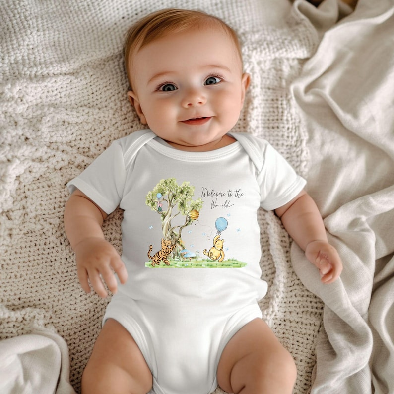 May include: A white baby onesie featuring a colorful illustration of Winnie the Pooh, Tigger, and a tree with balloons. The text "Welcome to the World" is written in script. The baby is lying on a cream-colored blanket.