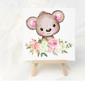 May include: A watercolor illustration of a brown bear with pink ears surrounded by pink and white flowers. The illustration is printed on a white card and displayed on a wooden easel.