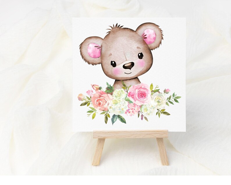May include: A watercolor illustration of a brown bear with pink ears surrounded by pink and white flowers. The illustration is printed on a white card and displayed on a wooden easel.