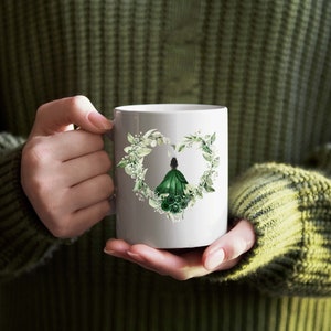 K&ouml;nnte beinhalten: Wei&szlig;e Keramiktasse mit gr&uuml;nem Blumendesign, das eine Frau in einem gr&uuml;nen Kleid zeigt. Das Design hat die Form eines Herzens.