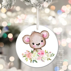 May include: A round white ceramic ornament with a brown bear with pink ears and a floral arrangement of pink and white flowers.