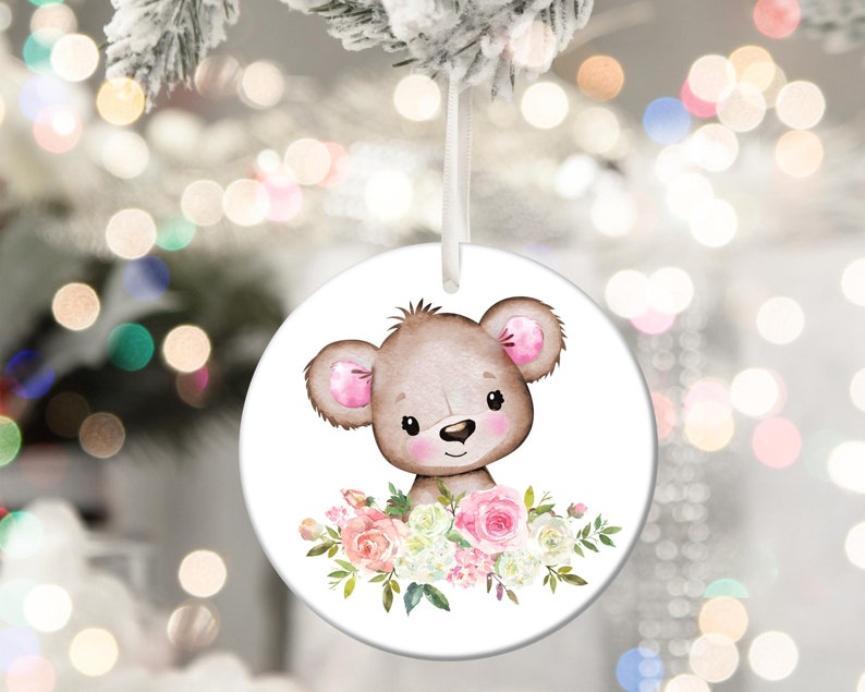 May include: A round white ceramic ornament with a brown bear with pink ears and a floral arrangement of pink and white flowers.
