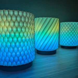 May include: Three 3D printed lamps with different patterns. The lamps are lit and glow in shades of blue and green.