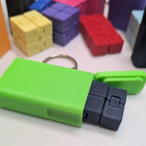 May include: A green keychain case with a black fidget cube inside. The case has a small opening to reveal the cube. The cube is made of plastic and has a textured surface.