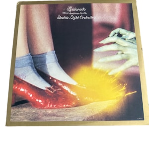 May include: A vintage vinyl record album cover featuring a pair of red shoes with a yellow flame effect. The album title is "Eldorado - A Symphony By The Electric Light Orchestra" in gold lettering.