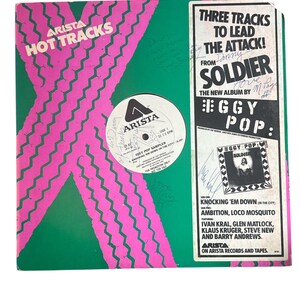 May include: A vintage vinyl record album cover with a green background and a pink zig-zag design. The album is titled "Iggy Pop Sampler" and features the text "Hot Tracks" and "Arista." The album cover also includes text about the album "Soldier."