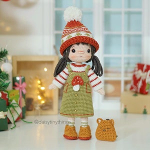 Pode incluir: Uma boneca de crochê vestindo um chapéu vermelho e branco com um pompom branco, um suéter listrado verde e branco, um vestido de macacão verde com uma aplicação de cogumelo e botas marrons. A boneca está segurando um pequeno animal de crochê marrom.