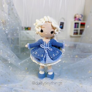 May include: A crocheted doll with long blonde hair wearing a blue dress and a blue cardigan. The doll is standing on a blue and white sparkly fabric.