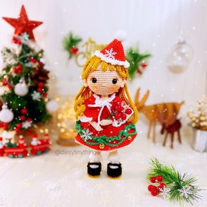 May include: A crocheted Christmas doll wearing a red dress with a green trim, a white snowflake hat, and holding a sprig of red berries. The doll has blonde hair and is standing on a snowy surface. The caption "@daisytinythings" is visible in the lower left corner of the image.