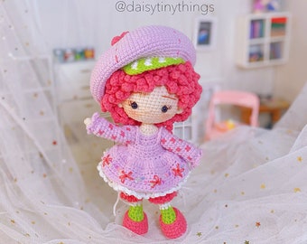 May include: A crocheted doll with pink hair, a pink and white dress, and a pink hat with green trim. The doll is standing on a white surface with gold stars.