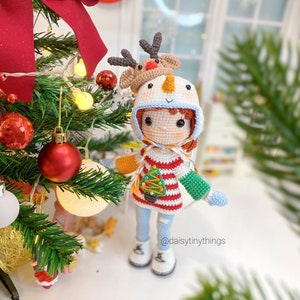 May include: A crocheted doll wearing a snowman hat with a reindeer on top. The doll is wearing a red and white striped sweater with a green Christmas tree on the front. The doll is holding a green and white crocheted mitten in one hand. The doll is standing in front of a Christmas tree with red and white ornaments.