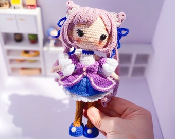 Könnte beinhalten: Eine gehäkelte Puppe mit rosa Haaren, blauen Augen und einem lila-blauen Kleid. Die Puppe trägt einen weißen Kragen und eine blaue Schleife. Die Puppe hält eine weiße Blume in der Hand. Die Puppe steht auf einer weißen Oberfläche. Der Text "Daisytinythings Handmadewithlove" ist im Hintergrund sichtbar.