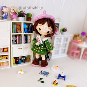 Puede incluir: Una muñeca de crochet con un gorro rosa y una chaqueta de cuadros verdes, sosteniendo un libro. La muñeca está de pie frente a una casa de muñecas en miniatura con estantes llenos de libros y otros artículos en miniatura.