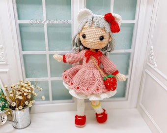 May include: A crocheted doll with a pink dress, white socks, and red shoes. The doll has a white headband with a red bow and a strawberry-shaped purse. The doll is standing in front of a window with white trim.