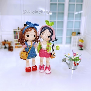 May include: Two handmade crochet dolls, each with unique outfits and accessories. One doll wears a blue denim jumper, a yellow purse, and red shoes. The other doll has a pink skirt, green jacket, and pink sneakers. Both dolls have dark hair and are holding hands.