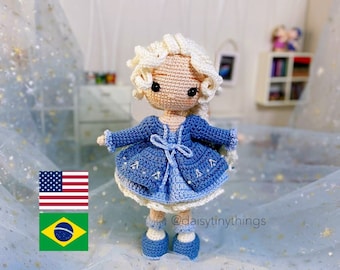 Könnte beinhalten: Eine gehäkelte Puppe mit blonden Haaren, die ein blaues Kleid und eine Jacke trägt. Die Puppe steht auf einem blauen und weißen Hintergrund mit einer US-amerikanischen und einer brasilianischen Flagge im Vordergrund. Der Instagram-Handle der Puppe ist @daisytinythings.