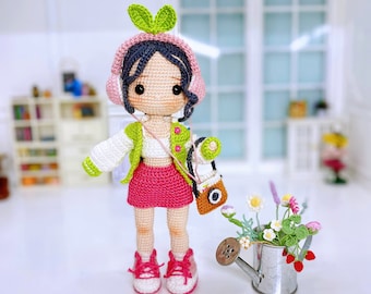 May include: A crocheted doll wearing a green jacket, pink skirt, and pink headphones. The doll is holding a small brown bag and is standing on a white surface. The doll has a green leaf on its head.