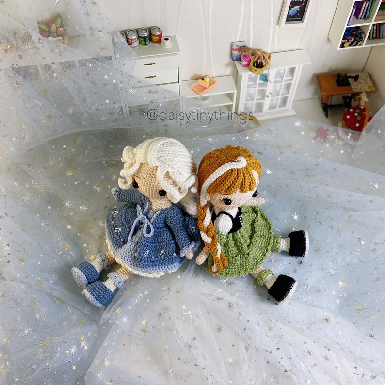 May include: Two crocheted dolls sitting on a white sparkly surface. The doll on the left is wearing a blue dress and has white hair. The doll on the right is wearing a green dress and has brown hair. The dolls are both looking to the right. The text "@daisytinythings" is visible in the image.