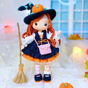 May include: A crocheted witch doll wearing a black hat with an orange pumpkin on top, a white and blue dress, pink shoes, and pink and white striped stockings. The doll is holding a small pink purse and is standing on a white surface with a broom behind her.
