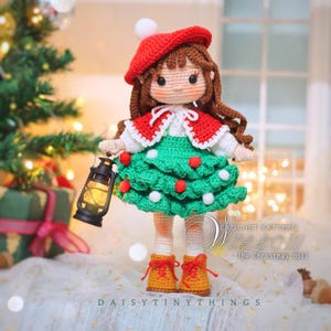 May include: A handmade crochet doll dressed for Christmas, wearing a red beret and cape over a green dress adorned with red and white accents. The doll holds a miniature lantern and is wearing yellow boots. The text "DAISY TINY THINGS" is visible.