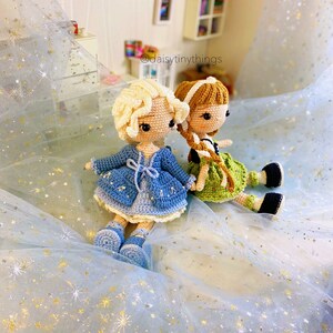 May include: Two crocheted dolls, one with blonde hair and a blue dress, the other with brown hair and a green dress, sitting on a blue sparkly background. The dolls have embroidered details and are holding hands. The text "@daisytinythings" is visible in the background.