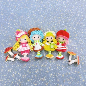 May include: Four handmade crochet dolls with different colored hair, hats, and outfits. Each doll has a unique design, including a strawberry, mushroom, and other decorative elements. Several crochet mushrooms are also present.