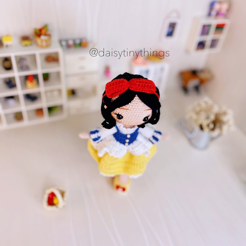 May include: A crocheted Snow White doll with a red bow, blue and white dress, and yellow skirt. The doll is standing on a white surface. The text "@daisytinythings" is visible in the background.