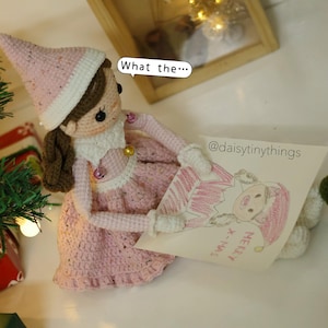 May include: A pink crocheted elf doll wearing a white hat with gold trim holds a drawing of another elf with the words "Merry Xmas" on it. The doll is sitting on a white surface with a Christmas tree in the background.