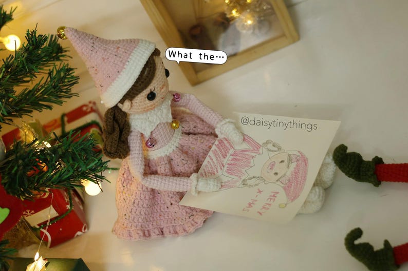 May include: A pink crocheted elf doll wearing a white hat with gold trim holds a drawing of another elf with the words "Merry Xmas" on it. The doll is sitting on a white surface with a Christmas tree in the background.