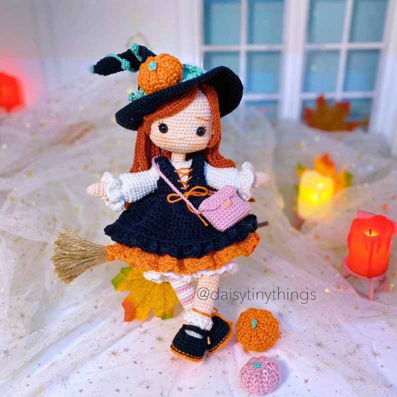 May include: A crocheted witch doll wearing a black dress with orange trim, a black hat with a green pumpkin on top, and a pink purse. The doll is holding a broom and is standing on a white surface with a few small pumpkins.