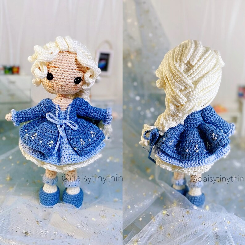 May include: A crocheted doll wearing a blue dress and white cardigan. The doll has long white hair in a braid and blue shoes. The doll is standing on a blue and white background with the text "@daisytinythin" on the bottom.